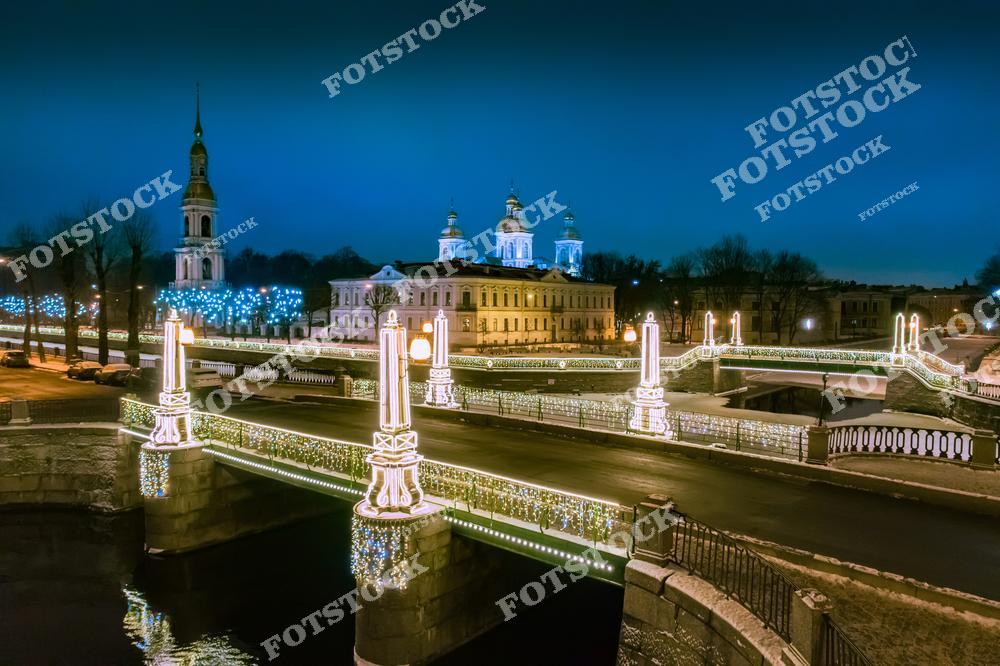 Фотография праздничного Санкт-Петербург, Россия, сделанная в новогодний период. На изображении запечатлены украшенные мосты через Крюков канал и Канал Грибоедова, подсвеченные яркой праздничной иллюминацией. Фотоснимок передаёт атмосферу Нового года в Петербурге: огни отражаются в воде, создавая торжественный и уютный облик зимнего города. В кадре также виден Никольский морской собор, подчёркивающий историческую и духовную часть городского пейзажа. Это изображение отражает красоту новогоднего Санкт-Петербурга и его знаменитые мосты и каналы.